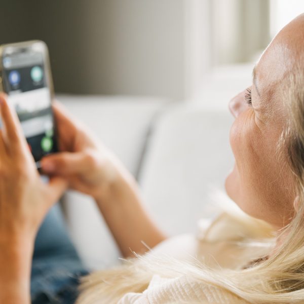 En dame med blondt hår som ligger på sofaen og smiler og holder en mobiltelen med en app-løsning fra porttelefon-leverandøren Urmet